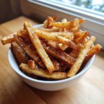 A close-up of a white bowl filled with golden brown, perfectly Crispy Air Fryer French Fries sprinkled with coarse salt.