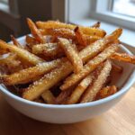 A close-up of golden brown, crispy air fryer french fries generously sprinkled with coarse salt in a white bowl.