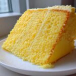 Close-up of a perfect slice of moist Yellow Velvet Cake with light frosting, served on a white plate near a window.