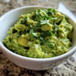 A close-up of chunky, bright green guacamole topped with fresh cilantro in a white bowl.