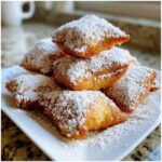 A stack of golden brown Vanilla French Beignets heavily dusted with white powdered sugar, served on a white plate.