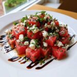 A vibrant Watermelon Salad featuring cubed watermelon topped with feta cheese, fresh mint, and a balsamic glaze drizzle.