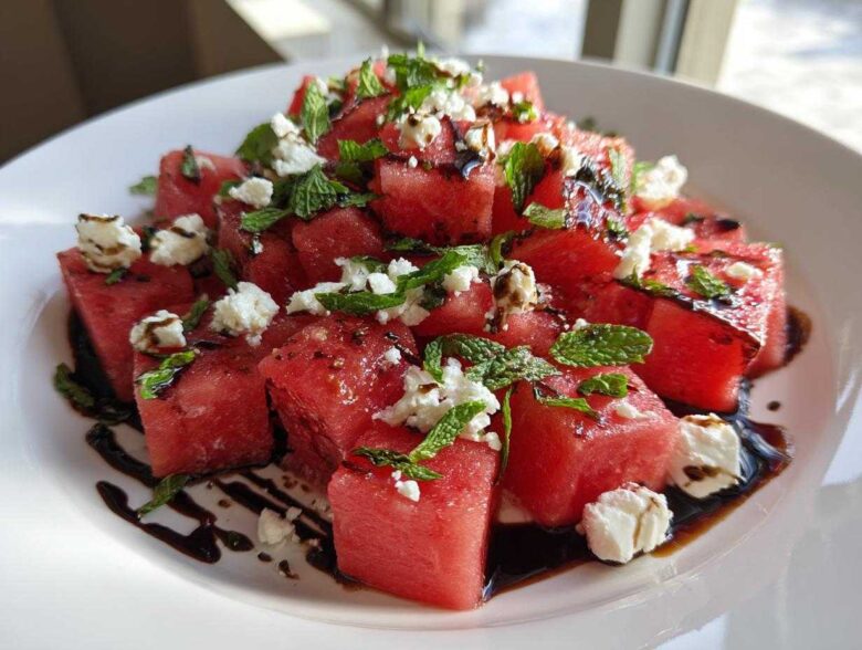 Close-up of a vibrant Watermelon Salad featuring cubed watermelon, crumbled feta cheese, fresh mint, and a balsamic glaze drizzle.